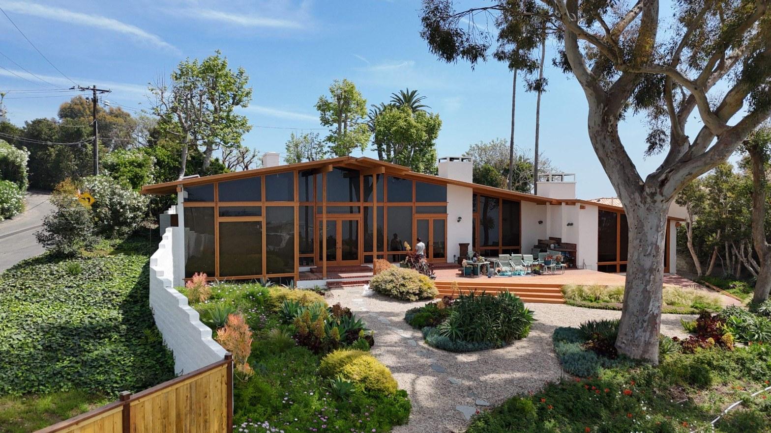 Mid-century modern single-story house with wood and glass walls, gravel garden, patio furniture, and mature eucalyptus tree.
