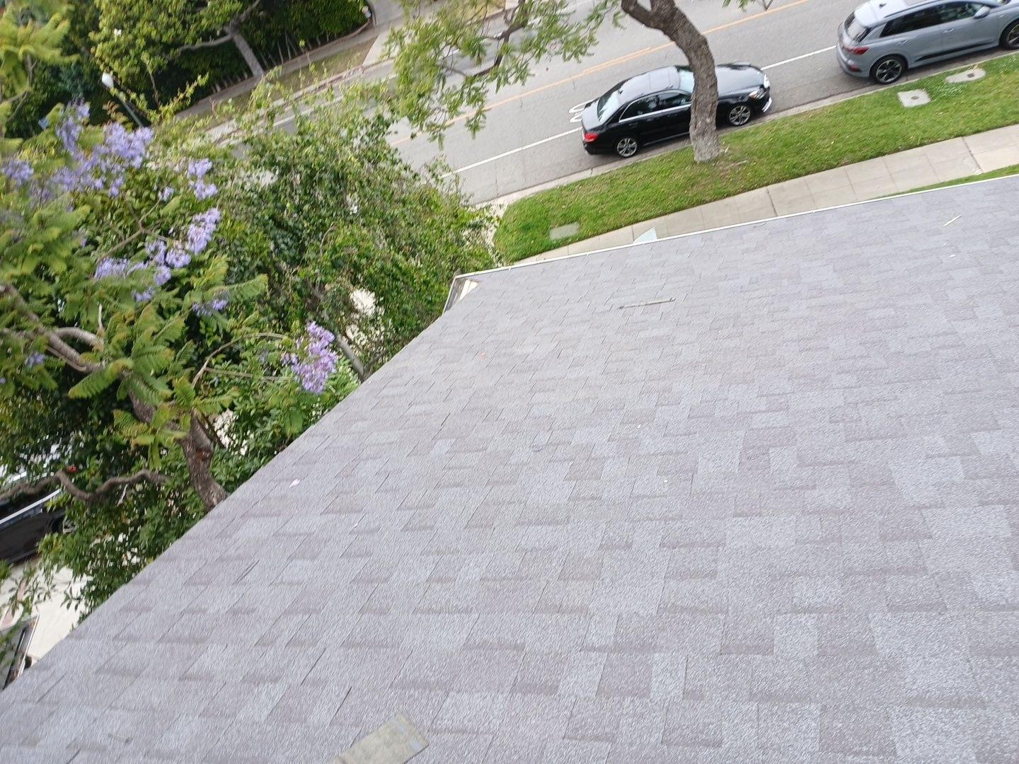 Overhead view of gray asphalt shingle roof on house with street, parked cars, and jacaranda tree visible below.