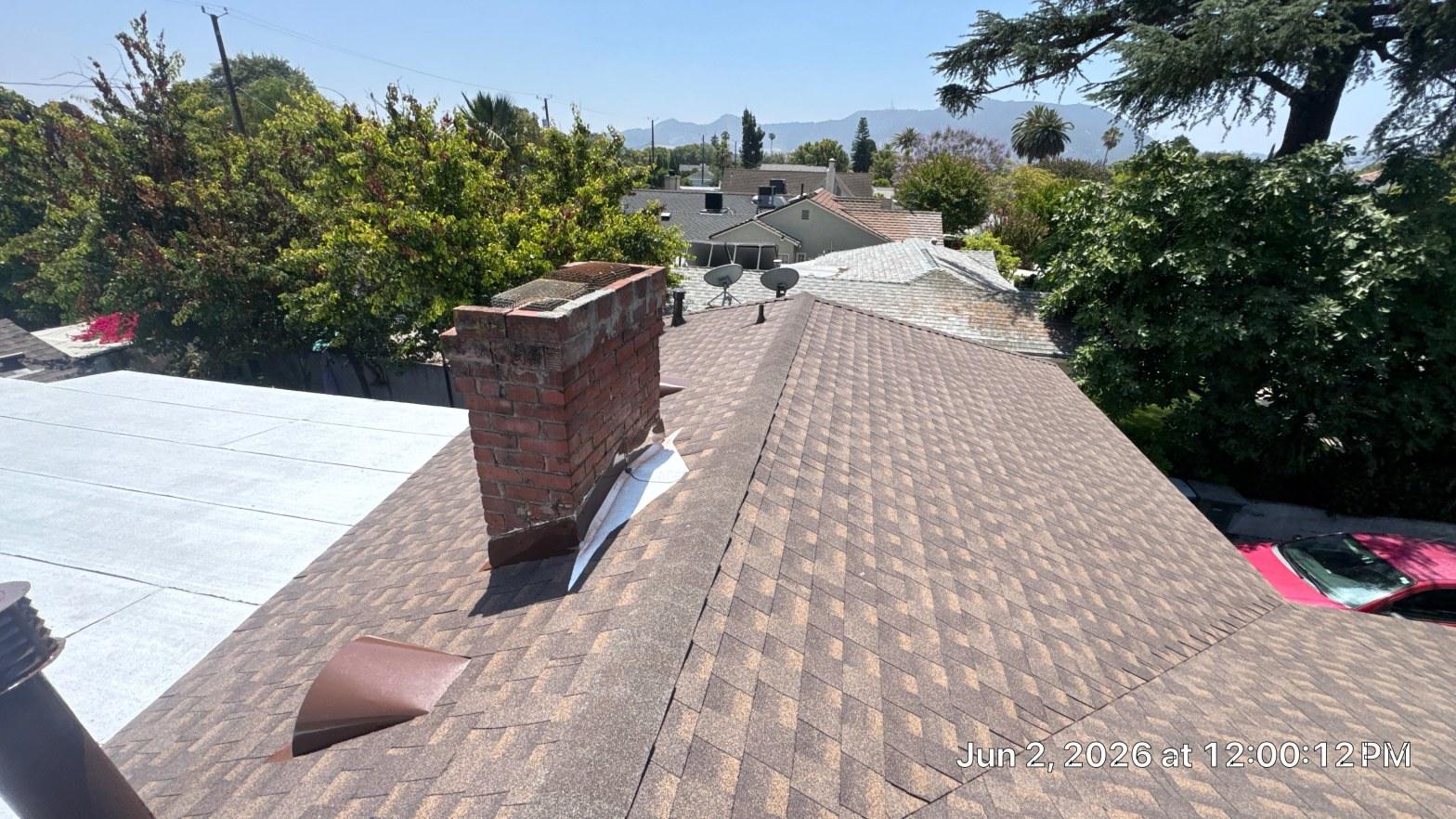 Deteriorating brick chimney with loose metal flashing on brown asphalt shingle residential roof, flat roof section adjacent.