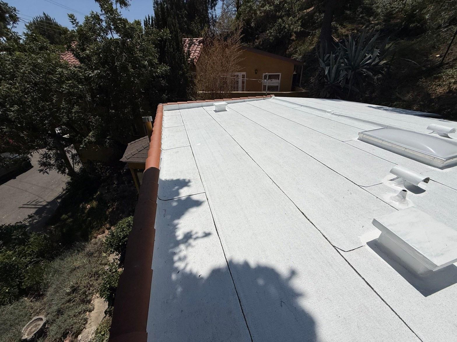 White flat roof coating with multiple skylights and red terracotta tile border on hillside home surrounded by trees.