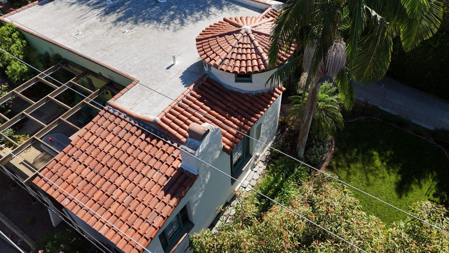Aerial drone photo of Spanish colonial home with terracotta barrel tile roofs, round turret, flat roof section, and palm trees.